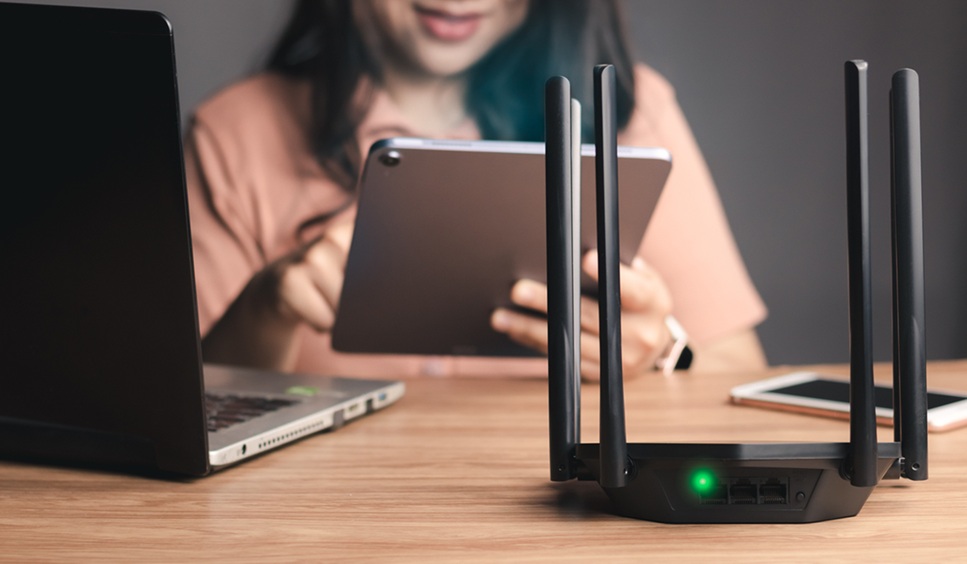 Selective focus at router. Internet router on working table with blurred happy woman using tablet at the background. Fast and high speed internet connection from fiber line with LAN cable connection. Selective focus at router. Internet router on working table with blurred happy woman using tablet at the background. Fast and high speed internet connection from fiber line with LAN cable connection.