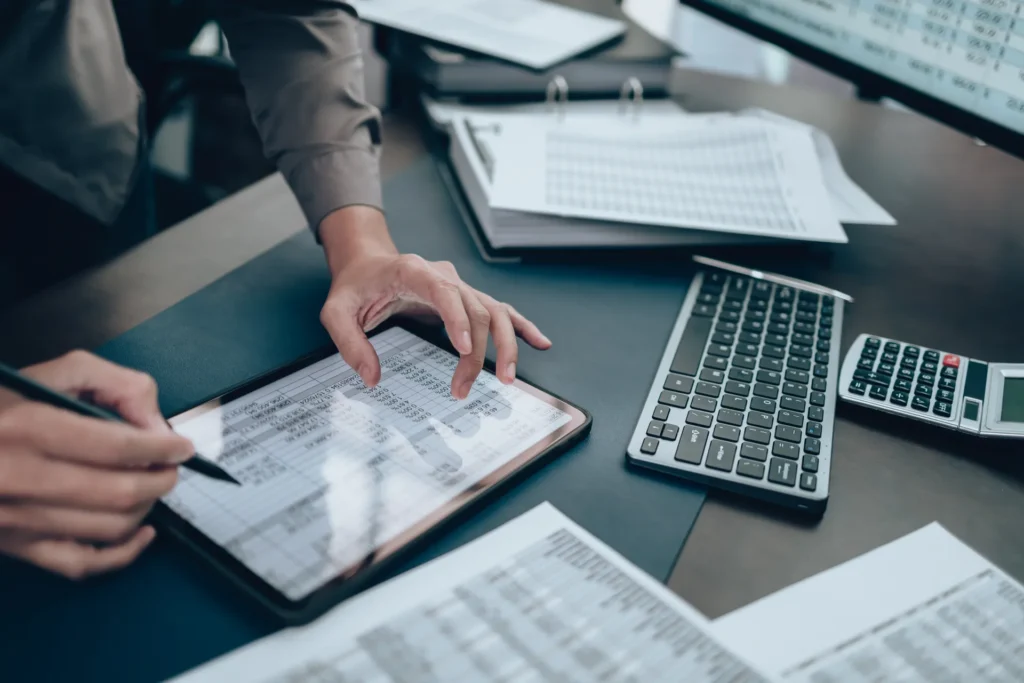 obrazek2_finance-tablet-arkusz-kalkulator-dokumenty-01 Osoba pracuje przy biurku, analizując dane finansowe na tablecie z arkuszem kalkulacyjnym, obok klawiatury, kalkulatora i papierowych dokumentów z tabelami.